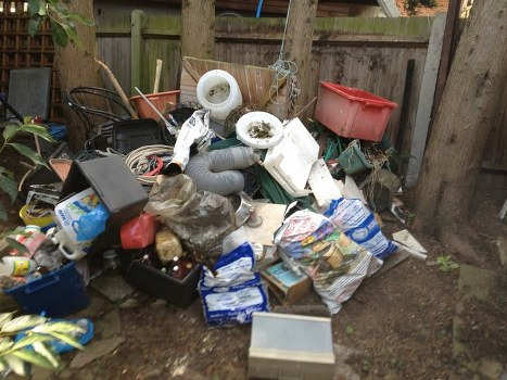 A pile of mixed household rubbish is gathered on a patch of dirt in a backyard, enclosed by a weathered wooden fence. The collection includes various materials such as white plastic pipes and fittings, a large red plastic storage bin, black and grey electronic cables, several paper and plastic packaging bags, and a cardboard box with miscellaneous small items. There are also plastic containers and bottles, some partially crushed, along with a black plastic tray and a small metal or plastic box. The items are stacked haphazardly, with some objects leaning against the wooden fence or lying on the ground. In the background, a few green leafy trees are visible over the fence. The scene is naturally lit, indicating daytime, and captures an outdoor environment suitable for rubbish removal services from Business Waste Removal Shoreditch, serving areas in east London.