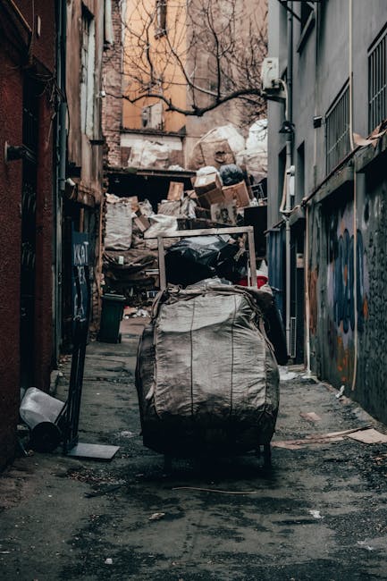 A narrow urban alleyway filled with accumulated waste, dominated by a large, gray, heavy-duty rubbish bag in the foreground, which appears to be made of durable plastic material with visible creases a